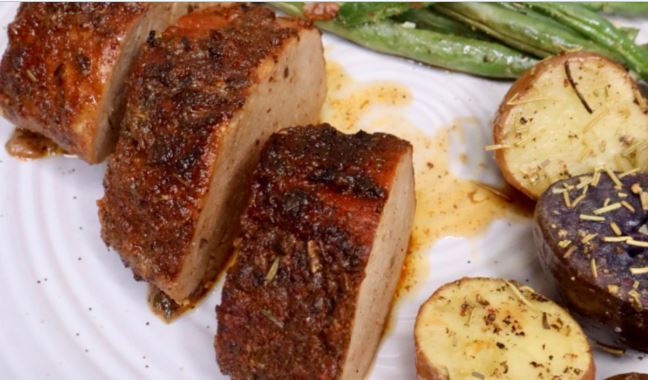 A perfectly cooked pork tenderloin sliced on a plate, topped with a rich sauce and garnished with fresh rosemary sprigs, next to a side of mashed potatoes and steamed vegetables.